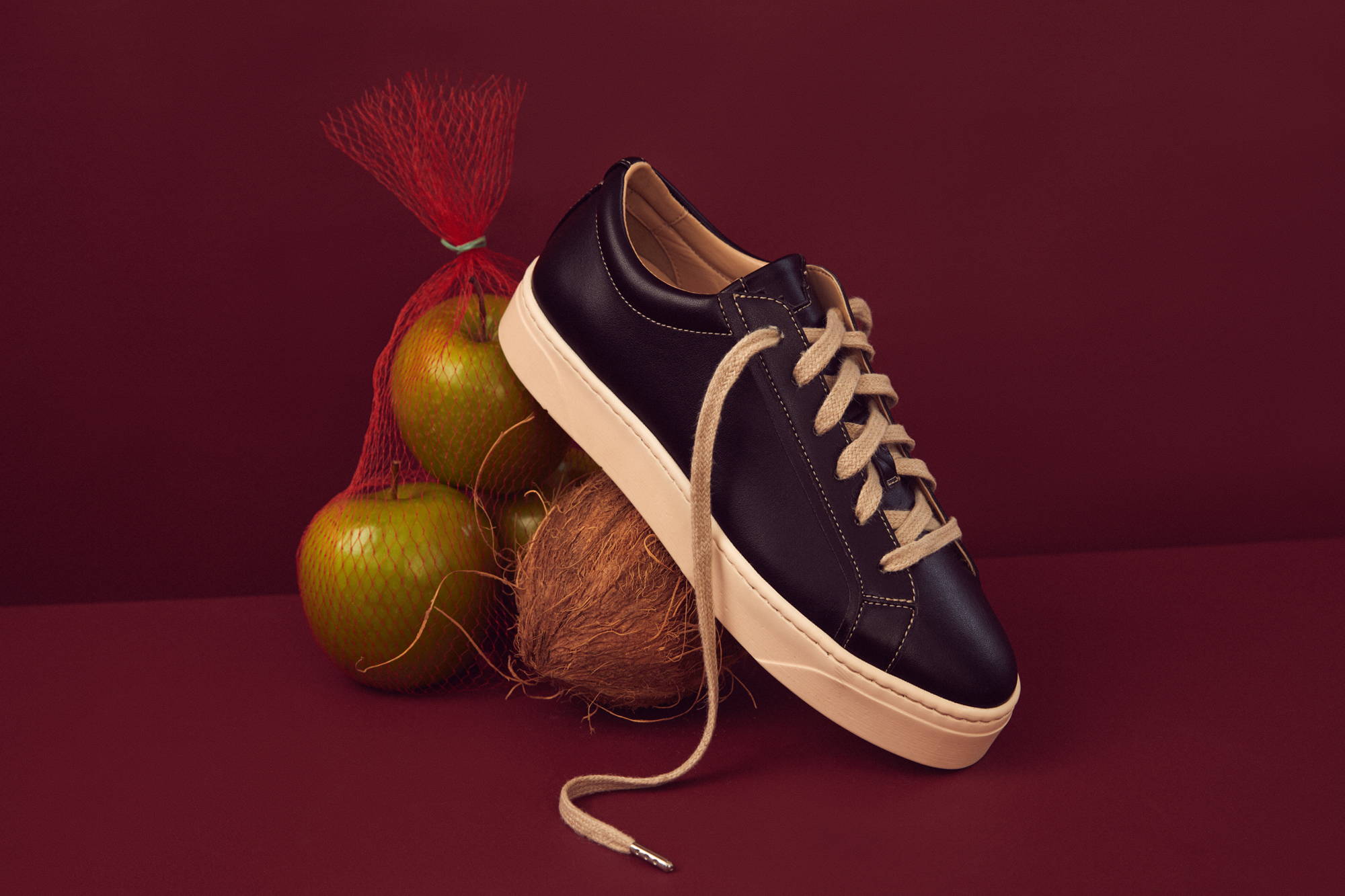 Plant-based luxury footwear - our Mel vegan apple and coconut sneaker pictured on a dark red background alongside apples and coconuts